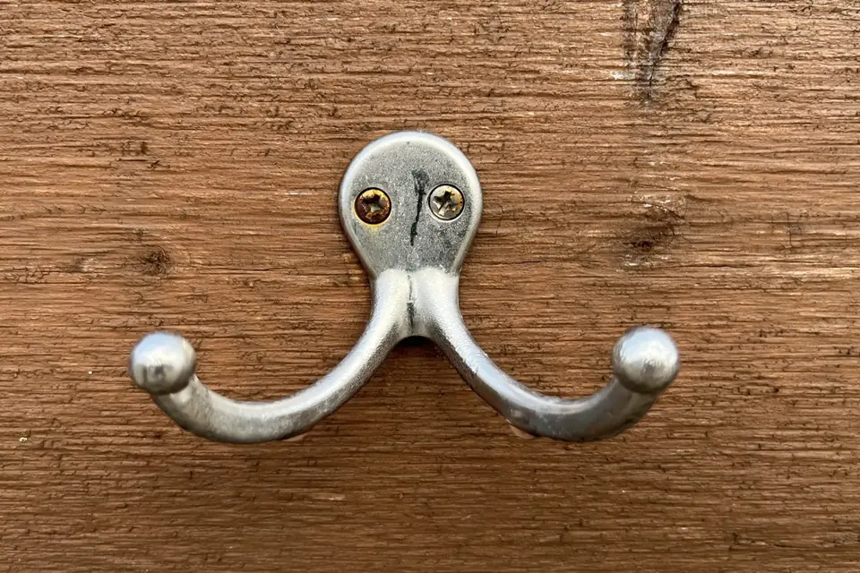 A double metal coat hook screwed into a weathered wooden board