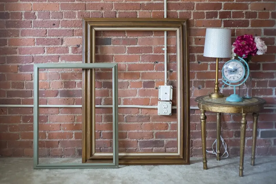 Empty picture frames leaning against a brick wall next to a small table with a lamp